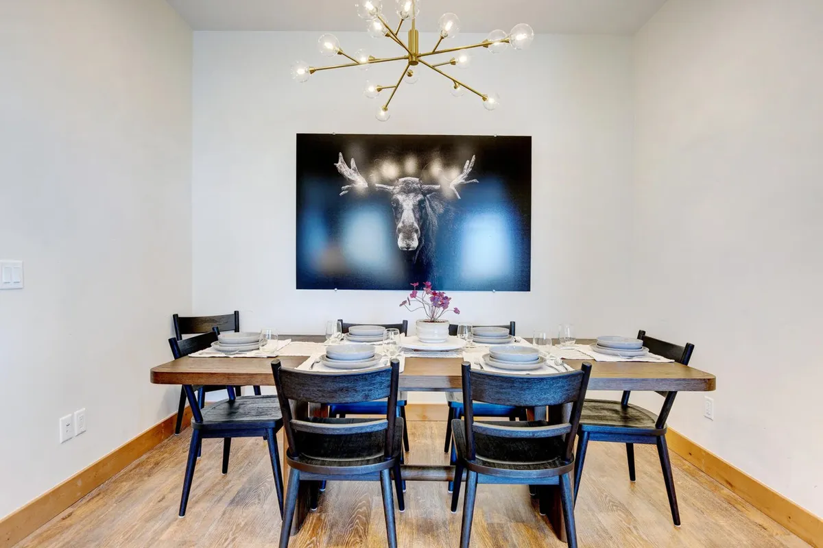 A spacious dining area featuring a large table that seats seven, illuminated by a striking chandelier. Artwork on the wall adds character and a welcoming, home-like touch. The space is designed for memorable meals and conversations, fitting for families and groups.