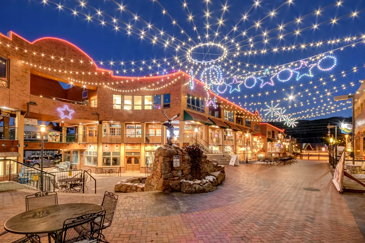 An inviting exterior scene in downtown Winter Park, illuminated by string lights and decorated with festive stars and snowflakes. The brick pathway winds past a stone fountain and outdoor gathering spaces, where people are seen strolling, shopping, and enjoying the atmosphere.