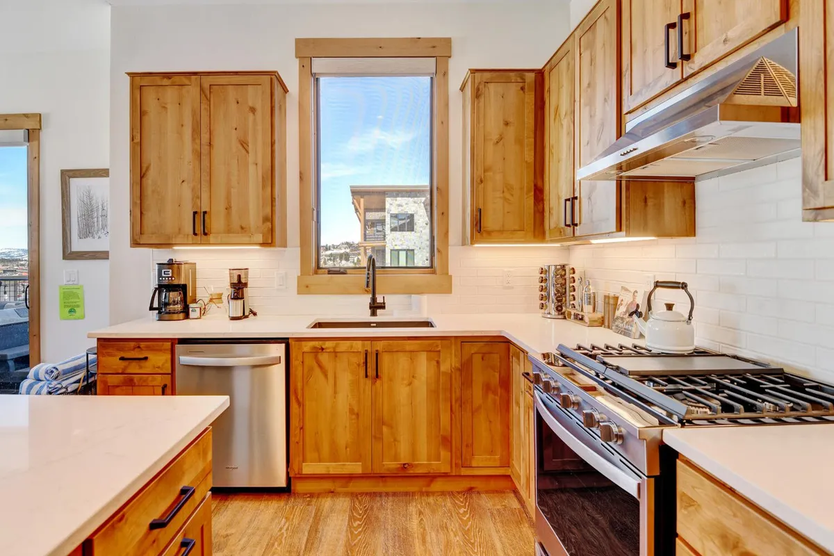 A modern kitchen bathed in natural light, featuring a large island, wooden cabinetry, stainless steel appliances, and a sizable window. The scene balances practical culinary space with a welcoming atmosphere for entertaining or casual meals.