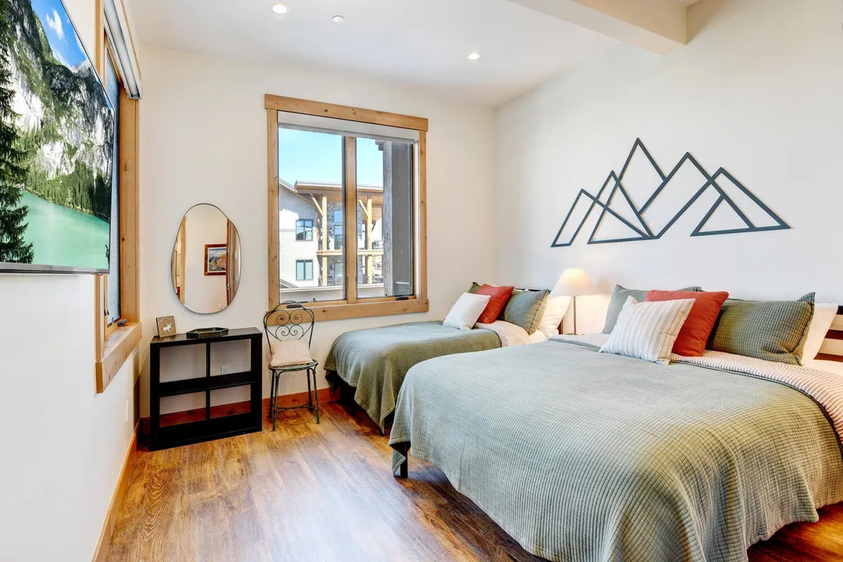 A welcoming second bedroom with two beds dressed in fresh green covers, mountain-themed wall art, a large window streaming in natural light, and a TV mounted on the wall. The space is adaptable for families or groups, encouraging shared moments and relaxation.