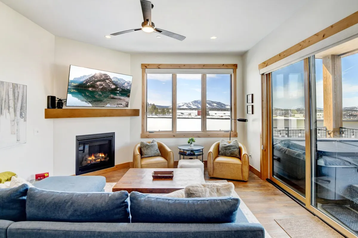 A modern living room with a comfortable blue sofa, a mounted television, a modern fireplace, and large windows framing expansive mountain vistas. The wooden coffee table and decor complete the luxurious yet cozy mountain atmosphere.