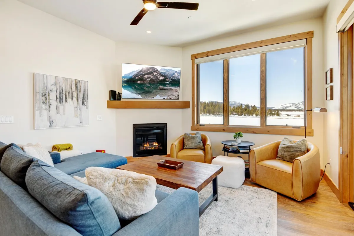 The image showcases a cozy living room with a large window framing breathtaking mountain views, a glowing fireplace, plush seating, and a TV mounted on the wall. The inviting setup suggests a perfect place to relax, unwind, or gather with loved ones while enjoying the natural beauty just outside.
