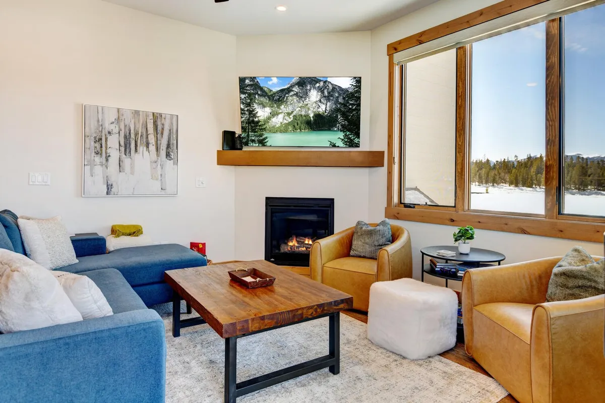 This image shows a comfortable seating area with plush furniture arranged around a coffee table. Large windows provide expansive views of the mountains, and a fireplace adds warmth and ambiance. The layout encourages relaxation and togetherness, while the natural light and mountain vistas bring the outdoors in.