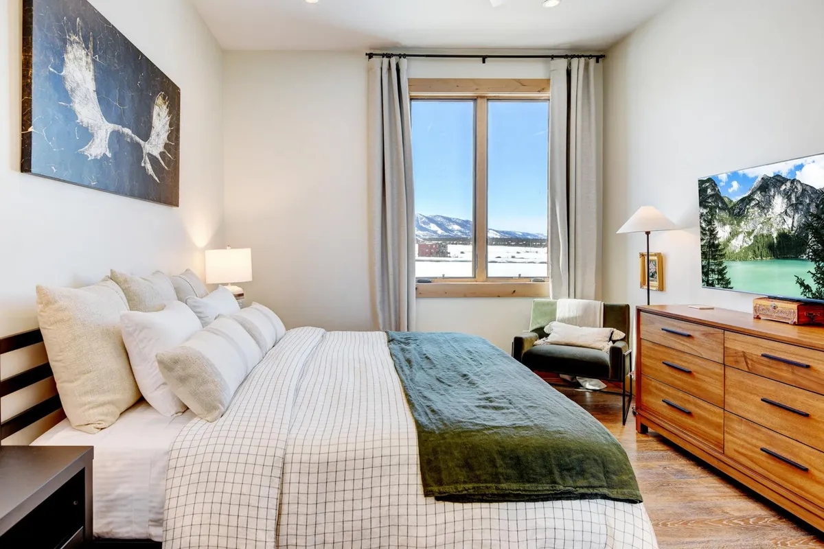 A serene bedroom featuring a comfortable bed with decorative pillows, a wooden dresser, artwork on the wall, and a large window offering breathtaking mountain views. The space exudes coziness and tranquility, with soft natural light filtering in.